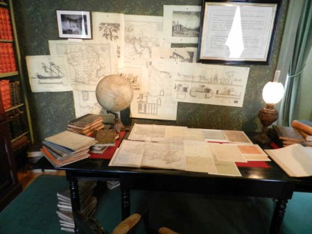 Dans la bibliothèque, une grande table est encombrée de cartes, de livres et d'un globe terrestre.